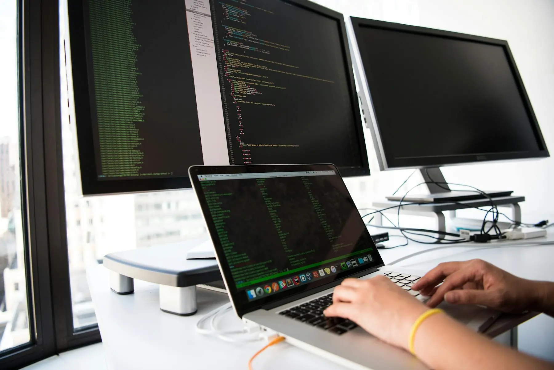 Bright software-development workstation with source code across a laptop and multiple monitors